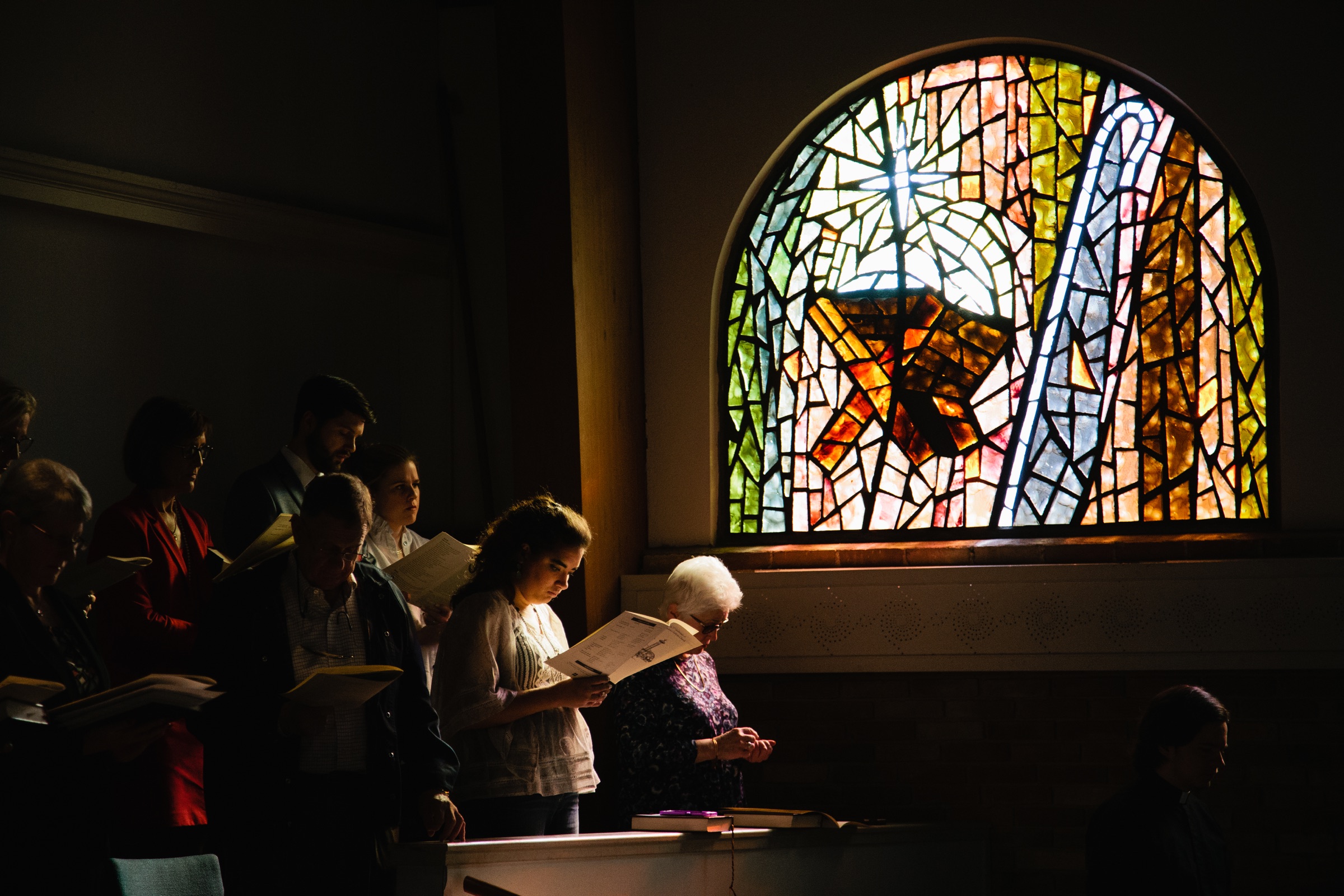Worshippers reading service sheets beside a sunlit stained-glass window.